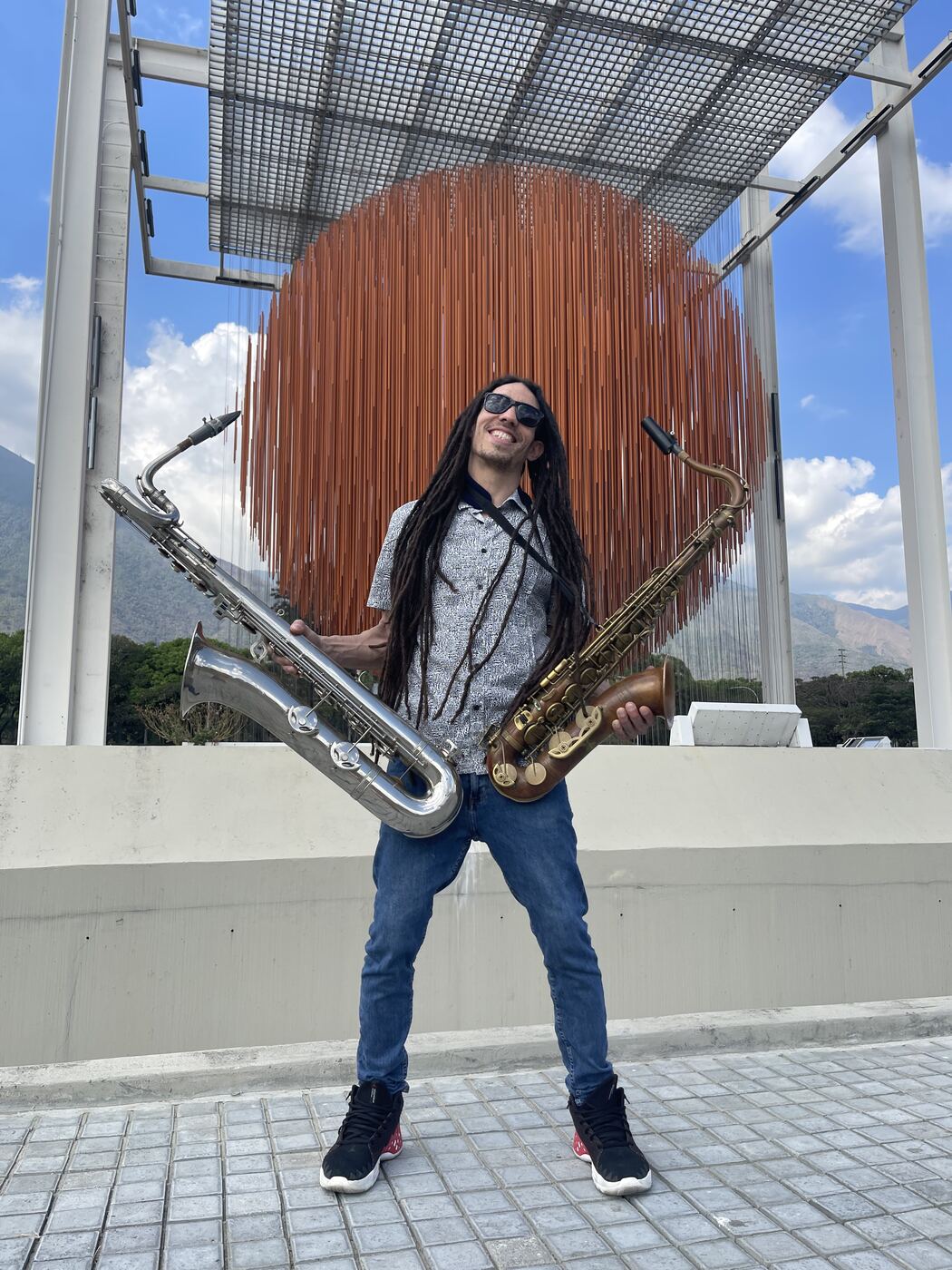 Kevin Alva smiling with a baritone and a tenor saxophone, one in each hand